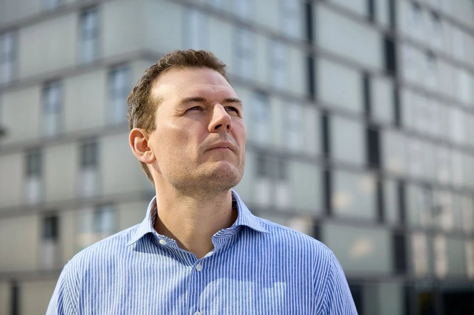 Man in blauw gestreept overhemd kijkt omhoog, voor een flatgebouw